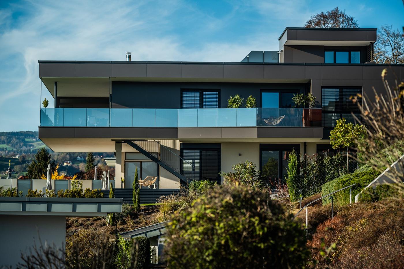 Wohnhaus am Hang mit dunkler Plattenfassade, auskragendem Obergeschoß und durchgehendem Glasbalkon